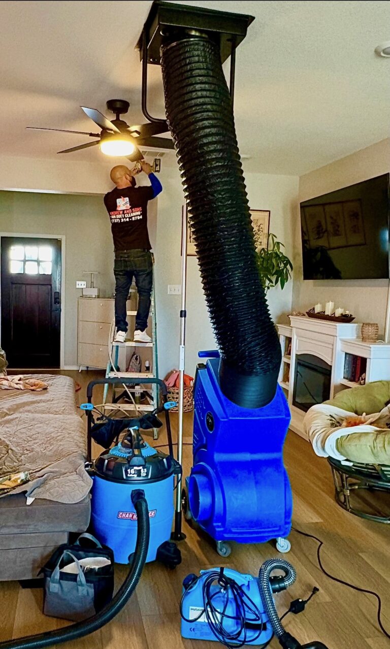 Andrew and Sons technician on a ladder cleaning a ceiling vent, with a negative air machine hose connected, in an Austin living room